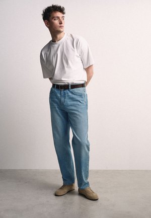 Jonge man staand met handen achter de rug, draagt een wit T-shirt, lichtblauwe spijkerbroek, bruine riem en beige schoenen, tegen een effen achtergrond.