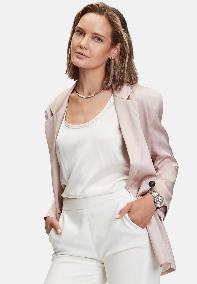 Helles pinkes Satin-Blazer über einem weißen Seidentop und cremefarbenen Hosen. Silberne Uhr mit Mesh-Armband. Kurze, zerzauste Haare und minimaler Schmuck.