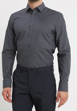 Homme portant une chemise ajustée à manches longues gris foncé boutonnée, rentrée dans un pantalon sombre avec une ceinture noire, debout devant un fond gris.
