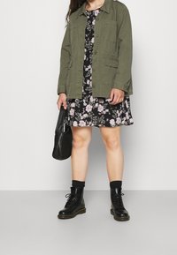 Olivgrüne Jacke mit Knopfverschluss, kombiniert mit einem schwarzen Blumenkleid. Schwarze Schnürstiefel und eine strukturiert schwarze Tasche runden das Outfit ab.
