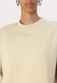 Sudadera beige con cuello redondo, hecha de una suave tela texturizada. Presenta un sutil logo bordado de "Calvin Klein" en el pecho.