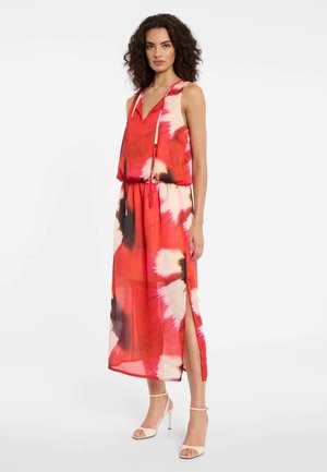 Femme aux cheveux bouclés portant une robe midi sans manches tie-dye rouge, blanche et noire avec une fente latérale et des sandales beige à talons hauts.