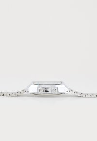 Montre argentée avec un boîtier rectangulaire, affichage digital et un bracelet tissé gris texturé. Comprend deux boutons sur le côté.