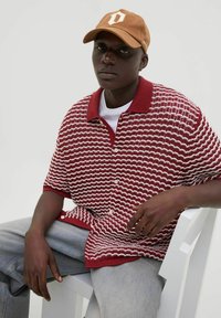 Polo-shirt tricoté à rayures rouges et blanches avec un col, assorti d'une casquette marron arborant un logo crème. Modèle assis sur une chaise blanche.