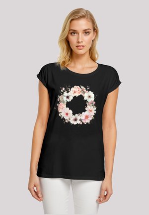 Schwarzes T-Shirt mit kurzen Ärmeln, das ein florales Kranzdesign in rosa und weißen Tönen zeigt. Hergestellt aus weichem Stoff mit entspanntem Schnitt.