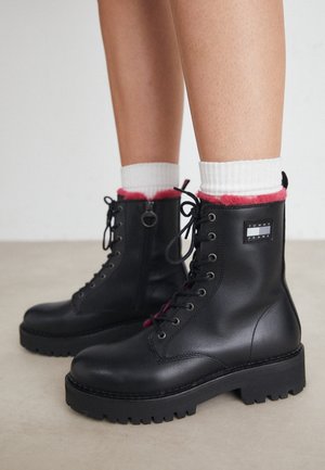 Czarne skórzane sznurowane botki na grubej podeszwie, z czerwonym wykończeniem i białymi skarpetkami widocznymi powyżej, noszone przez osobę stojącą na jasnej podłodze.