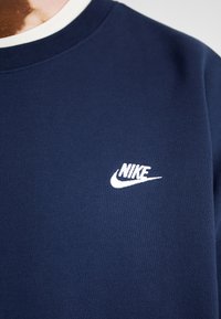 Sudadera azul marino hecha de un tejido suave, con un logo blanco de Nike en el área del pecho izquierdo y un cuello redondo con un ribete blanco en contraste.