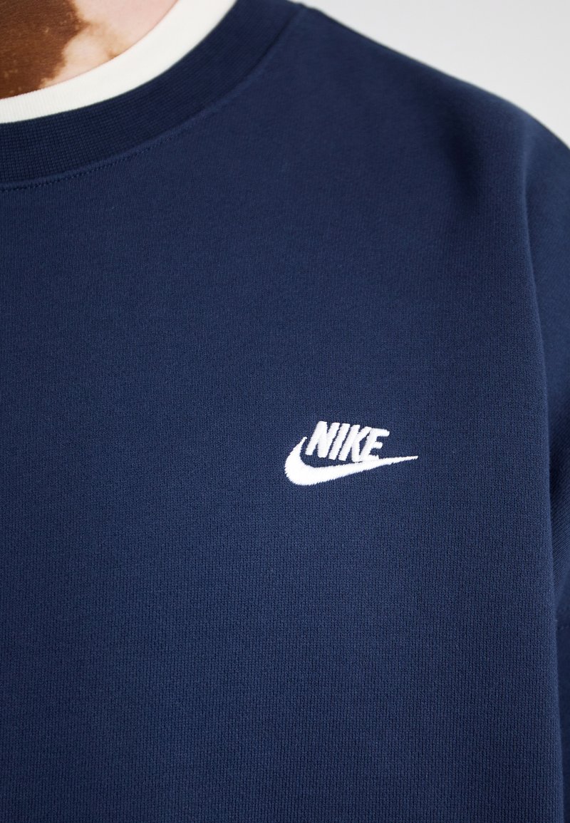 Sweatshirt bleu marine en tissu doux, avec un logo Nike blanc sur la partie gauche de la poitrine et un col rond avec un bord blanc contrastant.