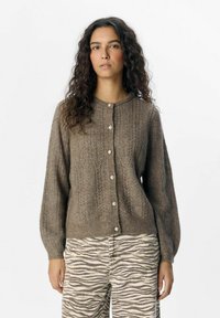 Cárdigan marrón de punto con cuello redondo, botones frontales y patrones texturizados. Mangas largas hechas de un hilo suave, combinadas con pantalones a rayas.