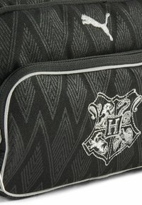 Bolsa de tela negra con un patrón geométrico texturizado, un logo de Puma en blanco y un escudo bordado con una 'H' destacada y diseños de animales.