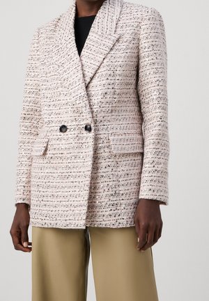 Person trägt ein hellrosa strukturiertes, doppelt geknöpftes Sakko mit schwarzen Knöpfen und beige Hosen mit weitem Bein.