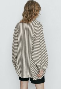 Luźna, paskowana bluzka w odcieniach beżu i brązu, z balonowymi rękawami i teksturowanym materiałem. Pokazana z tyłu, w połączeniu z ciemnymi szortami.