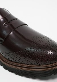 Mocassin en cuir brun foncé avec des détails perforés sur le bout, une lanière à fente et une semelle en caoutchouc épaisse et texturée.