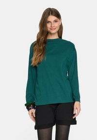 T-shirt z długim rękawem w kolorze teal, wykonany z mi ękkiej bawełny. Luźny krój z okrągłym dekoltem. Połączony z czarnymi szortami i wieloma kolorowymi bransoletkami.