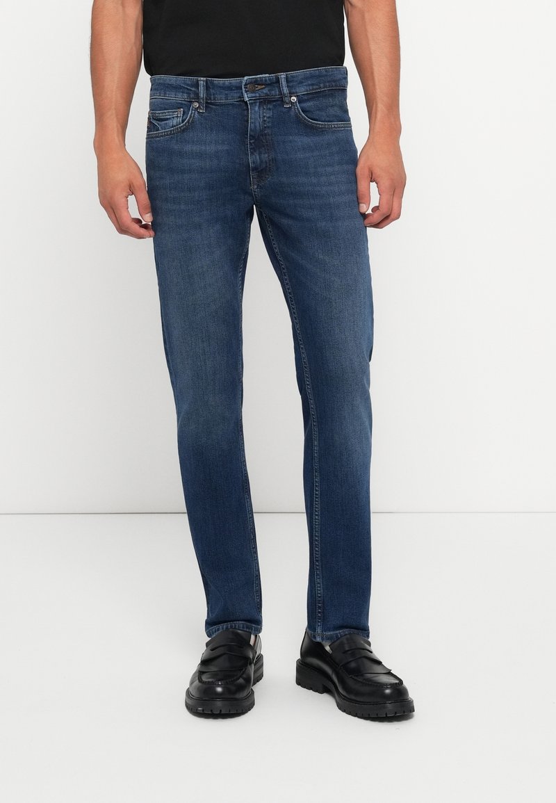 Homme portant un jean slim bleu foncé et des mocassins en cuir noirs, debout devant un fond blanc.