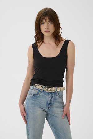 Femme aux cheveux bruns mi-longs, portant un haut noir sans manches, un jean bleu clair et une ceinture à motif peau de serpent.