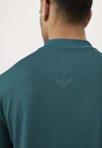 Moški nosi teal polo majico z embosirano logotipom orla, ki je centriran pod ovratnikom na hrbtni strani.