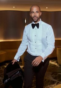Bărbat îmbrăcat cu o cămașă albă de tuxedo și cravată neagră, ținând o jachetă neagră, stând într-o cameră slab luminată, cu locuri curbe și podea din marmură.
