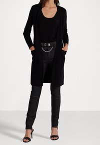 Svart lång kofta med öppen front, två sidofickor, ribbad struktur, kombinerad med en figurnära svart topp och tajta jeans, accentuerad med ett bälte.