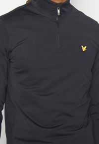 Svart långärmad polo med hög krage, försedd med en halvmeterdragkedja och en liten gul fågel-logotyp på bröstet. Sl ät textur, lättviktsmaterial.