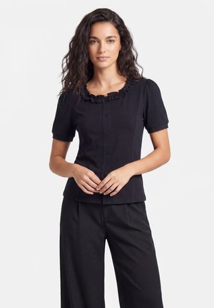 Femme aux longs cheveux ondulés porte un chemisier noir à manches courtes avec col à volants et boutons, assorti d’un pantalon noir, face à l’avant.