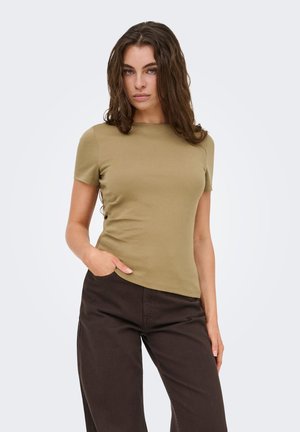 Femme aux longs cheveux bruns portant un haut vert olive à manches courtes et un pantalon marron foncé, une main dans la poche, sur un fond uni.