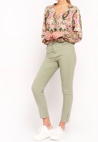 Vloeiende, gemusterde blouse in roze, groen en beige; gecombineerd met aansluitende lichtgroene broeken met een rits bij de enkel en witte sneakers.