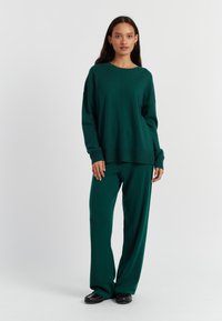 Pull en tricot vert et pantalon ample assorti. Texture douce, manches longues et décolleté large. Chaussures noires à semelle en caoutchouc.