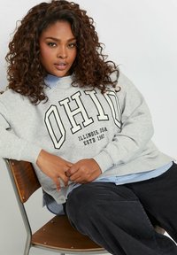 Felpa grigia con grafica "OHIO", indossata sopra una camicia a righe blu e bianche. Pantaloni scuri e una sedia in legno completano l'outfit casual.