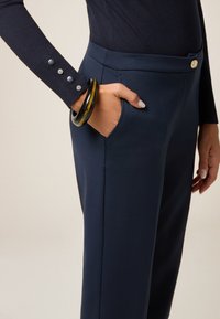Pantaloni navy con una trama liscia, dotati di una tasca laterale e di un bottone dorato. La manica ha tre bottoni argentati come accento.