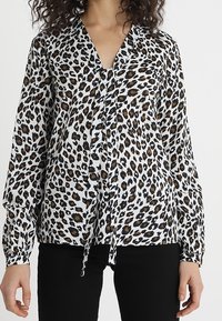 Leopardskinnad blus i vitt, svart och brunt, med långa ärmar och en knytframsdetailj. Tillverkad av mjukt, lätt tyg.