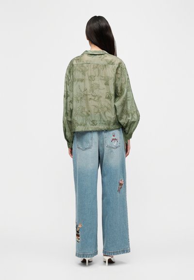 Διαφανές μπουφάν σε ελιά πράσινο με κουμπιά και ανάγλυφα λουλούδια· ψηλόμεσα ανοιχτό μπλε τζιν με ζωντανή κεντητική στα πόδια.