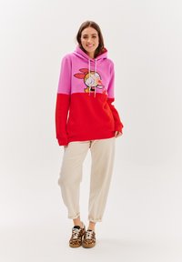 Barevná mikina s růžovými a červenými sekcemi, se směšnou postavou grafika. Doplňena béžovými kalhotami a botami s tygrovaným vzorem.