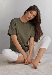 Oliveově zelené bavlněné tričko s krátkým rukávem, kulatým výstřihem a volnějším střihem, v kombinaci s bílými zkrácenými kalhotami na světlém texturovaném povrchu.