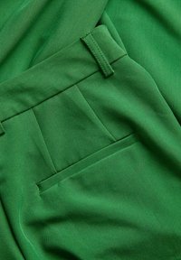 Groene broek met een gladde stoftextuur, een vlakke voorkant met plooien en een achterzak. De tailleband heeft riemlussen voor een goede pasvorm.