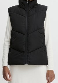 Czarna pikowana kamizelka z wysokim kołnierzem, pionowym zamkiem błyskawicznym i pikowanym wzorem. Noszona na jasnym, długim rękawie.