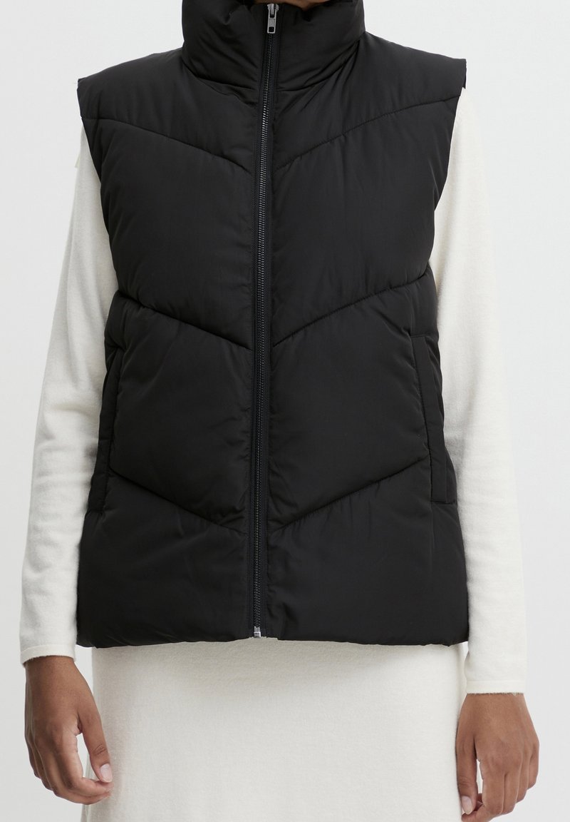 Czarna pikowana kamizelka z wysokim kołnierzem, pionowym zamkiem błyskawicznym i pikowanym wzorem. Noszona na jasnym, długim rękawie.