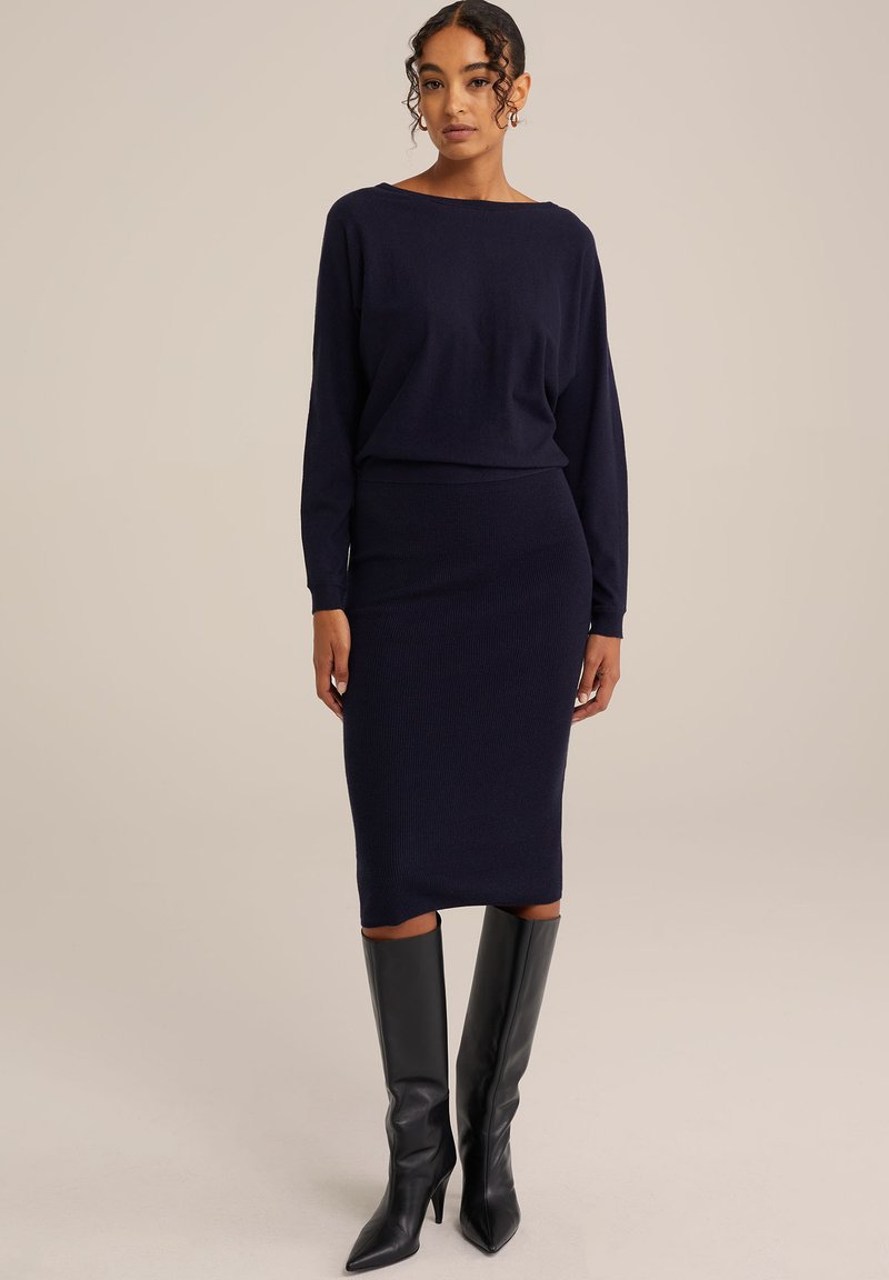 Navy gestrickter Pullover mit weitem Ausschnitt, kombiniert mit einem figurbetonten navy Midi-Rock und kniehohen schwarzen Lederstiefeln. Glatte Textur und minimalistisches Design.