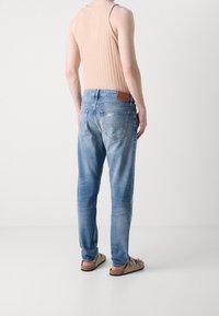 Ljusblå denimjeans med rak passform, blekta sektioner och synliga sömmar. Brun läderlapp på baksidan av midjan. Bruna sandaler bärs.