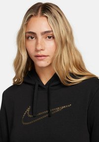 Černá mikina s kapucí, na níž je vyšívané zlaté logo Nike. Má stahovací kapuci a jemnou texturu. Detailní záběr na oděv na modelce.