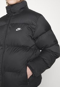 Sort dunjakke med høj krave, der har en glansfuld overflade, syede sektioner og et lille hvidt Nike-logo på brystet. Lommer med lynlås.