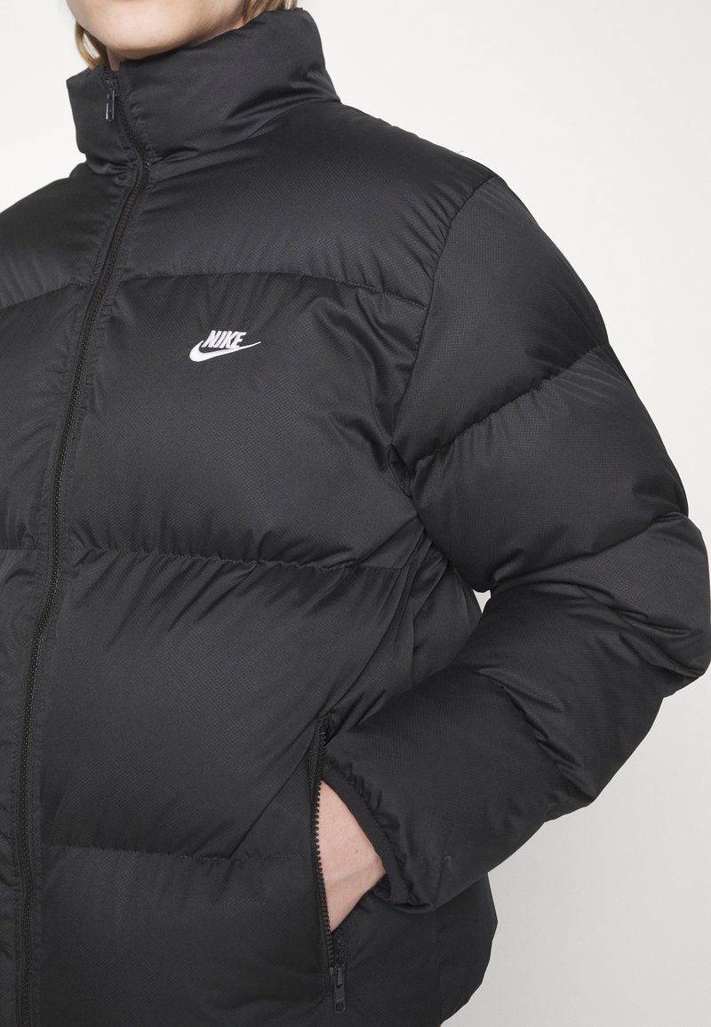 Chaqueta de plumas negra con alto cuello, con un acabado brillante, secciones cosidas y un pequeño logo blanco de Nike en el pecho. Bolsillos con cremallera.
