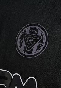 Okrągłe czarno-szare logo z rzymską cyfrą XXIII, kształtami geometrycznymi i małymi ikonami tarczy na teksturowanym czarnym tle z tkaniny.