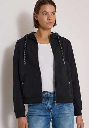 Femme portant un sweat à capuche noir zippé avec cordons sur un t-shirt blanc et un jean bleu, debout devant un fond clair uni.