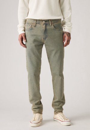 Mann trägt hellgrüne, ausgewaschene Jeans, weißes langärmliges Hemd und weiße Turnschuhe und steht vor einem schlichten Hintergrund.