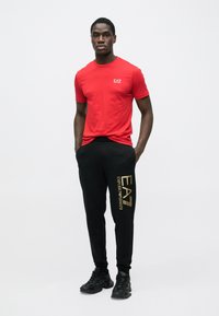 Maglietta rossa a maniche corte con logo sul petto abbinata a pantaloni della tuta neri con nome del marchio stampato in oro, indossata con scarpe da ginnastica nere.