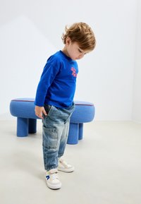 Blauw shirt met lange mouwen en rode afbeelding, lichtgewassen cargobroek, en beige sneakers met blauwe accenten. Ronde blauwe gestoffeerde kruk op de achtergrond.