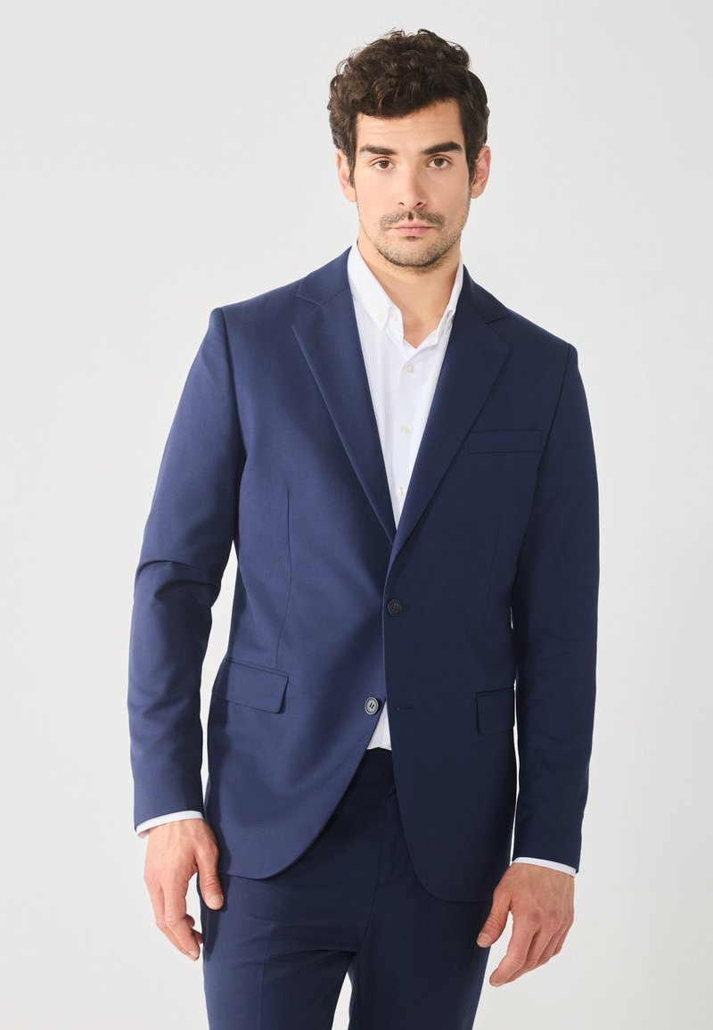 Blazer azul marino hecho de tela suave, con un cierre de un solo botón, solapas con muesca y bolsillos laterales; acompañado de una camisa blanca por debajo.