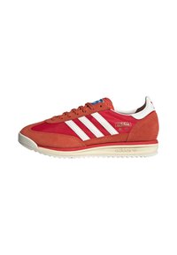 Zapatillas de ante rojo con franjas blancas, diseño retro. Presentan una suela de goma beige y acentos azules en la etiqueta de la lengüeta.