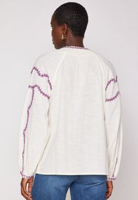 Frau mit Kurzhaar trägt eine weiße Bluse mit langen Ärmeln und lila gestickten Streifen sowie blauen Jeans, von hinten vor einem schlichten Hintergrund gezeigt.